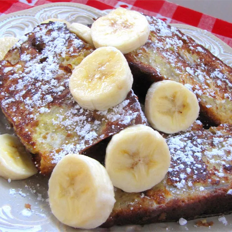 3 slices of banana bread french toast stacked on a white porcelain plate. Bread slices are dusted lightly with powdered sugar and topped with sliced banana coins about 1/2-inch thick.