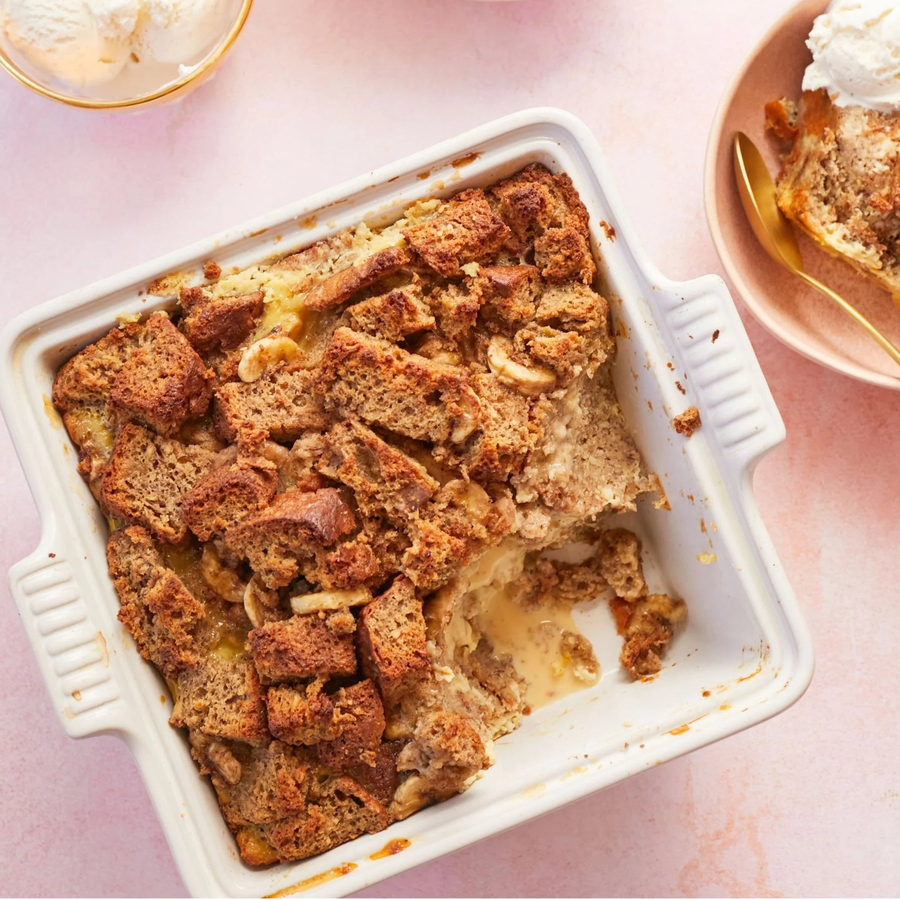 Baked Banana Bread Pudding in a white stoneware baking dish with handles. Photo is set on a white counter. Gold-trimmed clear bowl of vanilla ice cream i nthe top left corner. Beige colored bowl to the right of the baking dish filled with some of the banana pudding and a scoop of vanilla ice cream at the top of the dish. There is a brushed brass spoon in the bowl.