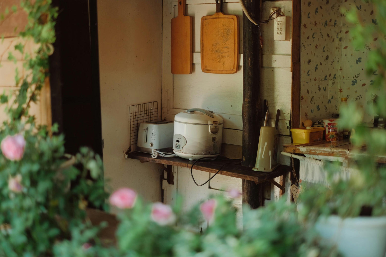 A cozy kitchen corner with vintage cookware, cutting boards, and floral wallpaper, partially framed by soft-focus greenery.