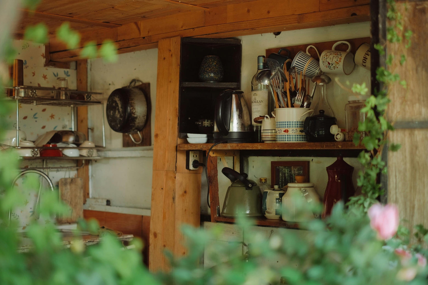 A rustic wooden kitchen shelf with teapots, mugs, and vintage cookware, framed by soft-focus greenery and pink blossoms.