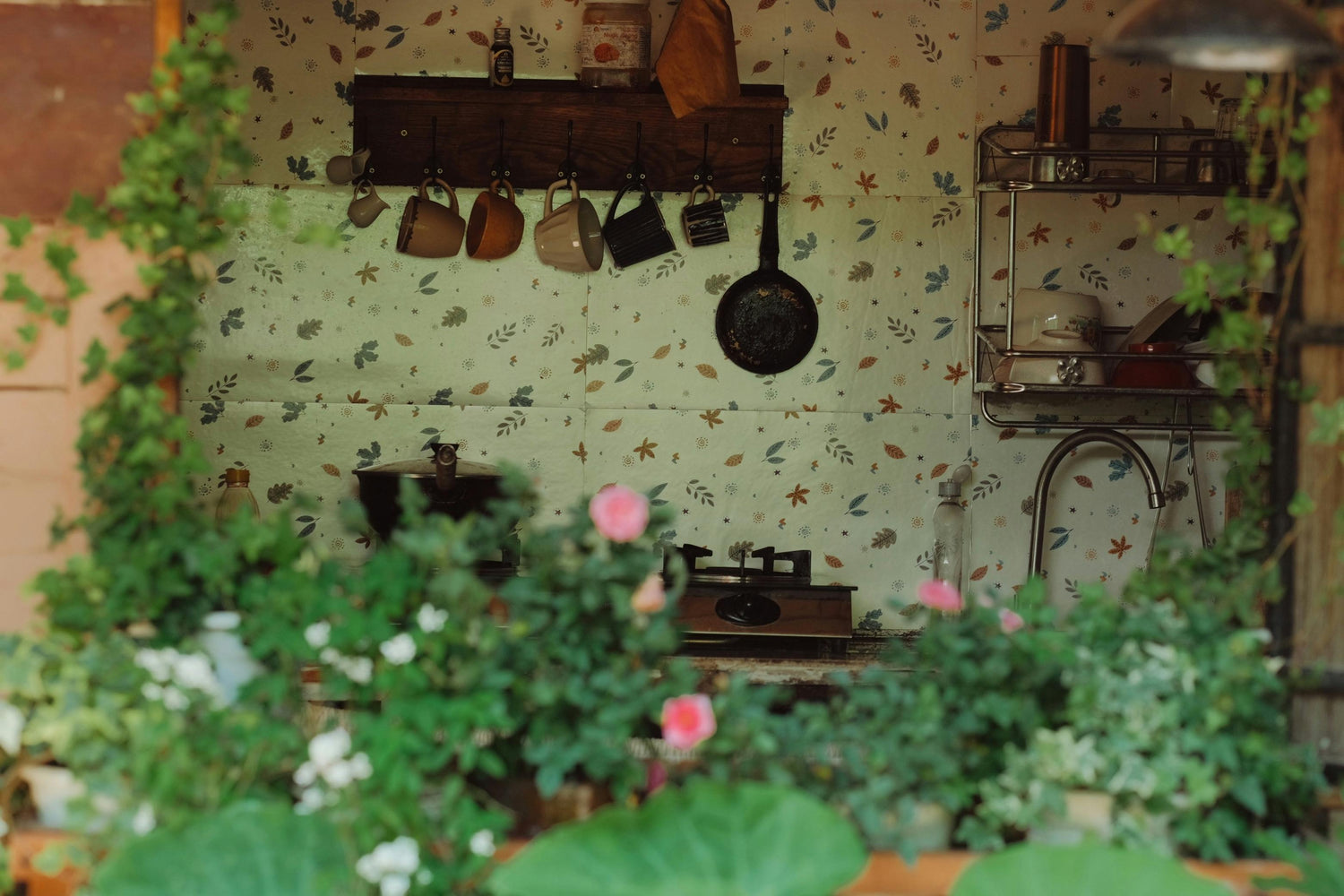 Vintage kitchen nook with floral wallpaper, hanging cookware, and open shelving, partially obscured by blooming greenery in the foreground.