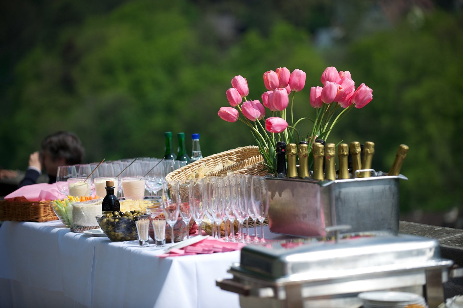 Outdoor catering setup featuring champagne bottles in ice, wine glasses, appetizers, and a vase of pink tulips, with greenery in the background.